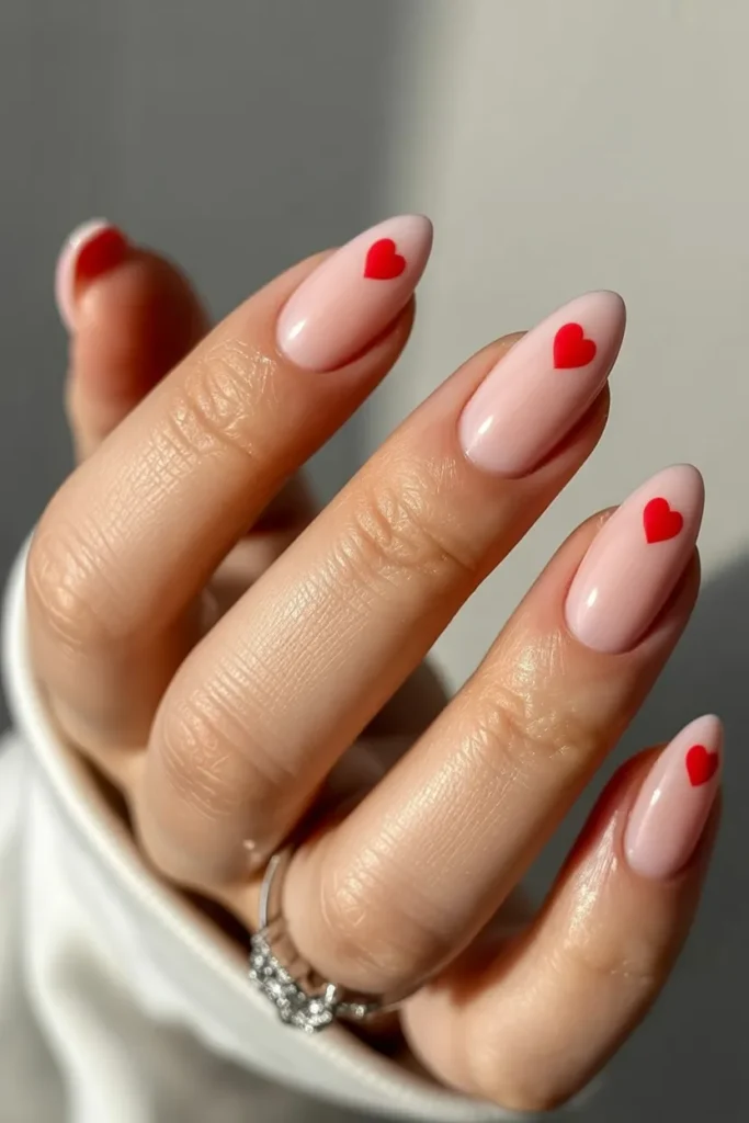 Close-up of medium-length almond-shaped nails painted nude beige with minimalist red heart accents, glossy salon finish, soft natural lighting, feminine hand wearing a silver ring, Valentine’s aesthetic trending nail art.