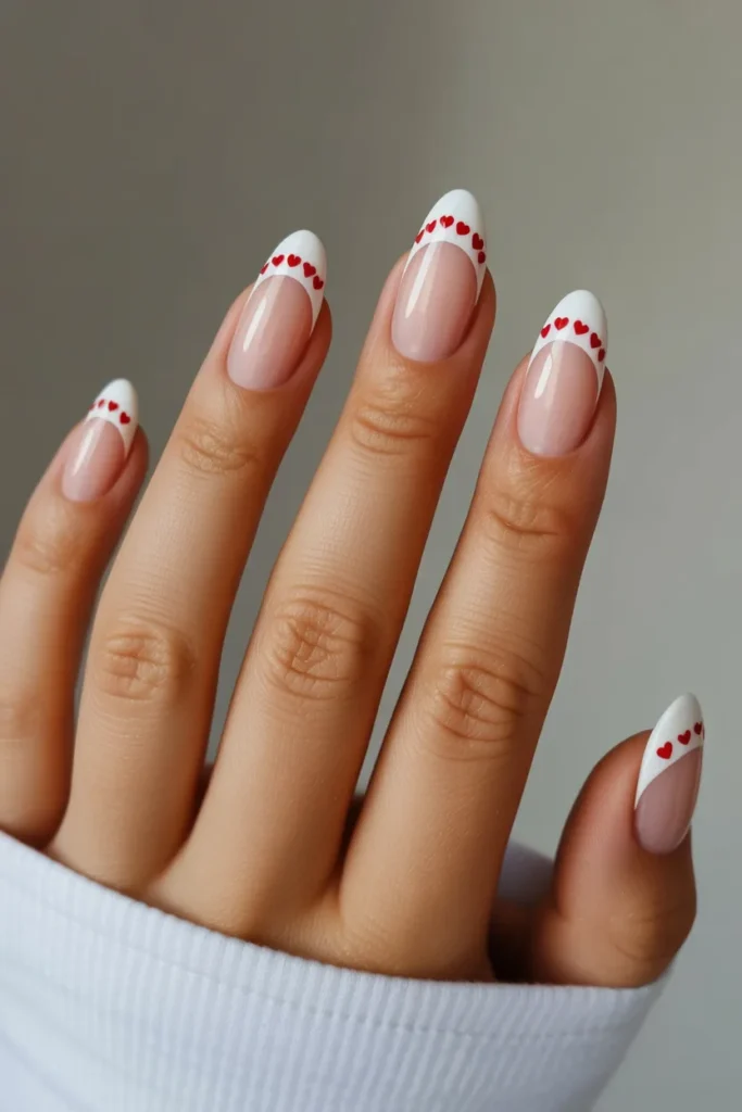 Medium almond nails with classic white French tips, small red hearts along the smile line, glossy professional finish, elegant modern Valentine’s nail design.